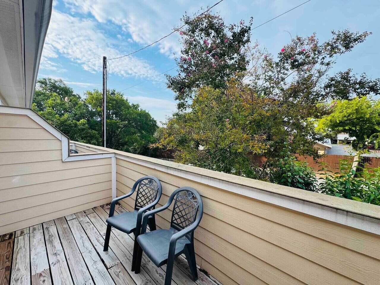 Terraza de madera con dos sillas y barandilla, con vista a un jardín con árboles y cielo despejado.