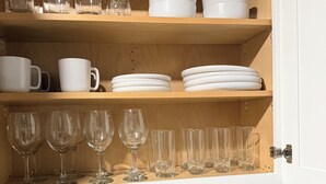 A kitchen with wooden shelves holding white cups, plates, and glasses. Below, there's a blender, a roll of paper towels, and a knife block.
