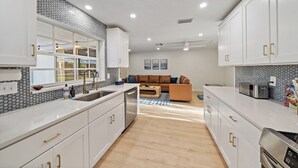 A modern kitchen with white cabinets, a dishwasher, and a microwave. There is a wooden dining table and a sofa in the adjacent living area.