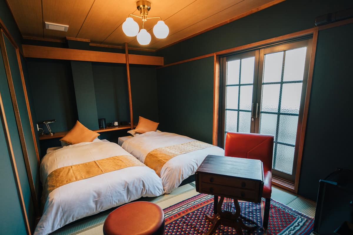 A hotel room with two beds, a wooden desk, a red chair, a patterned rug, and a large window with a view.