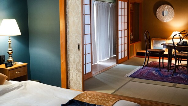 Una tradizionale stanza in stile giapponese con pavimento in tatami, un tavolino con sedie, un letto con biancheria bianca e porte scorrevoli che danno su un balcone.