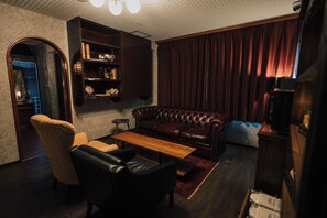 Un salon avec un canapé en cuir, une table basse en bois, un fauteuil à motifs, une bibliothèque et une grande fenêtre avec des rideaux.