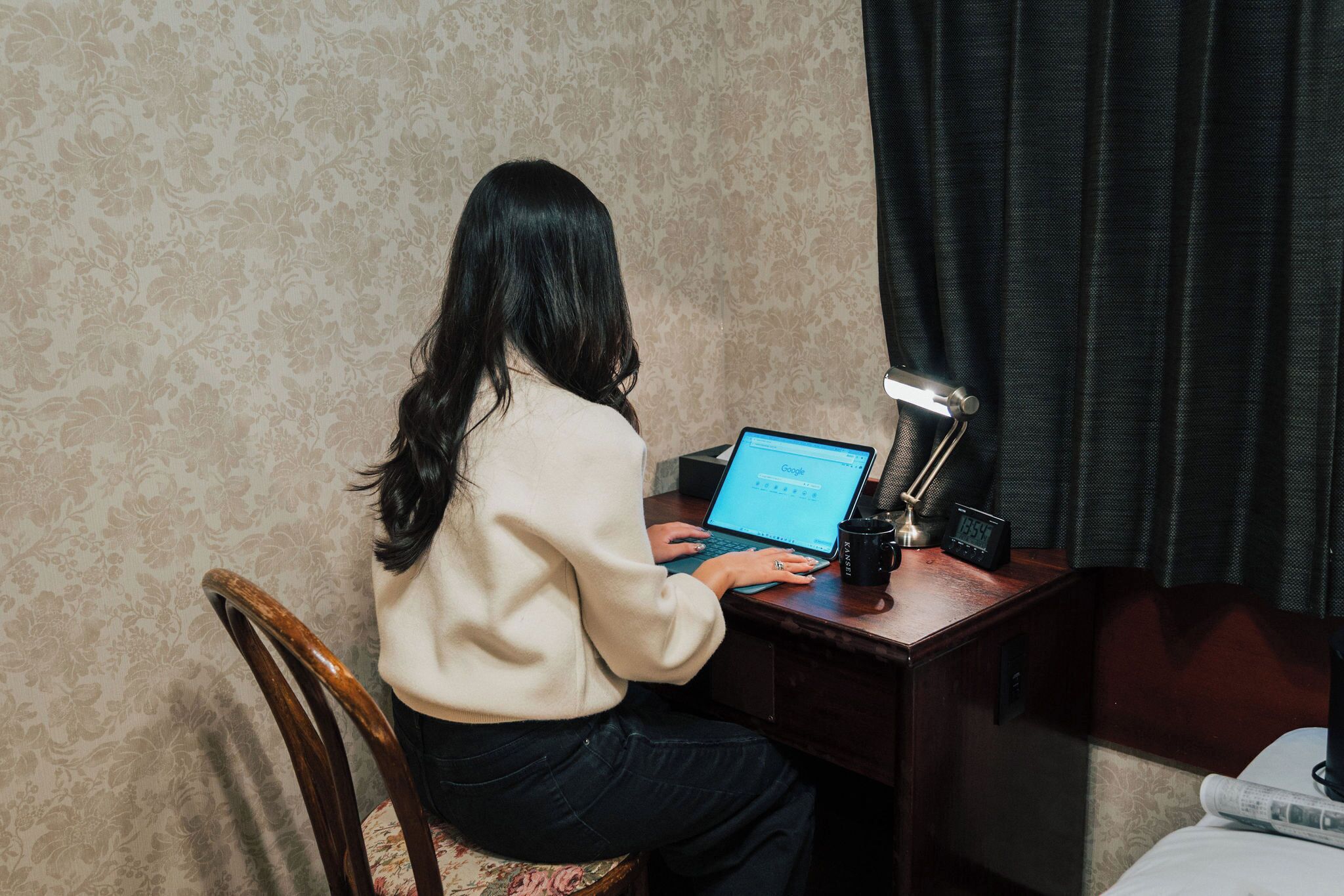 Uma pessoa sentada à mesa com um laptop, um relógio e uma luminária.