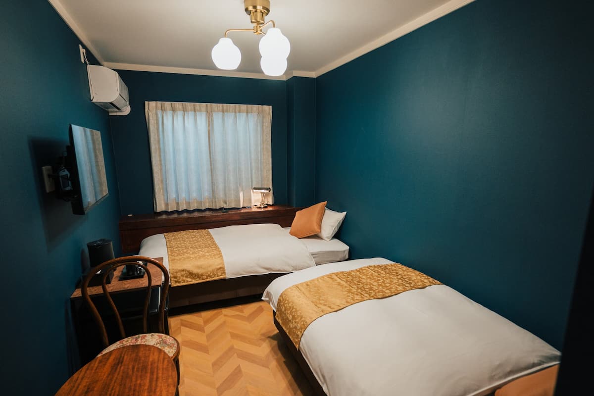 A hotel room with two beds, a desk, a chair, a window with curtains, and a ceiling light fixture.