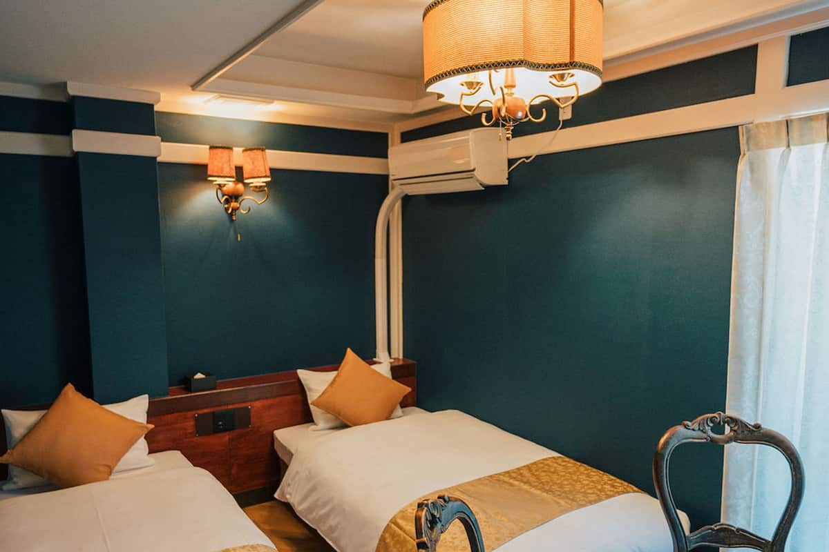 A hotel room with two beds, a wooden headboard, a chair, and a wall-mounted lamp.