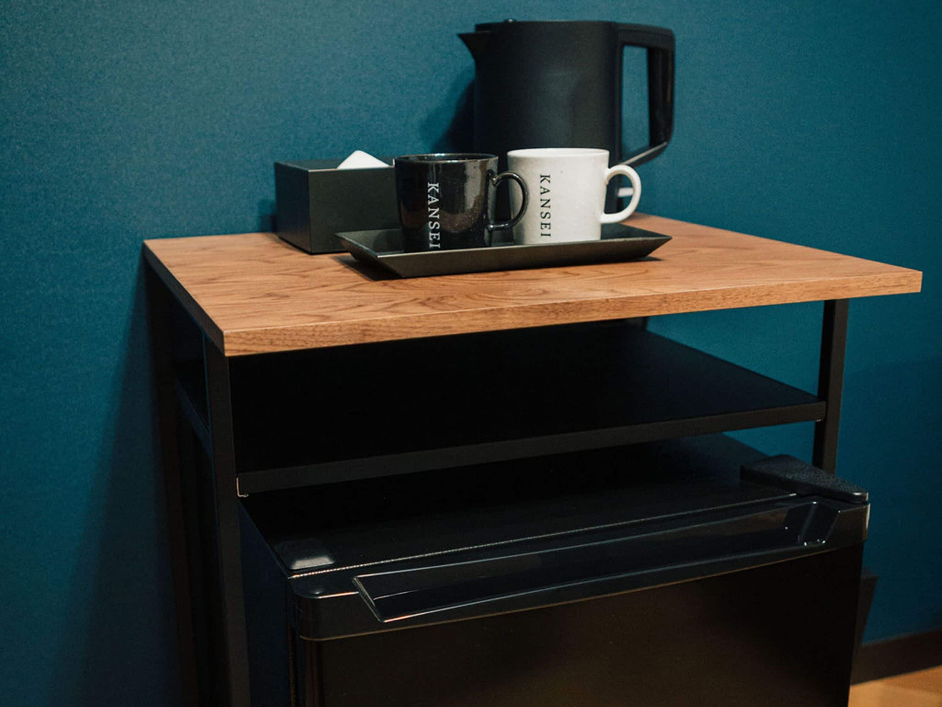 A small wooden table with a black tray holding a black coffee maker, a white mug, and a black box.