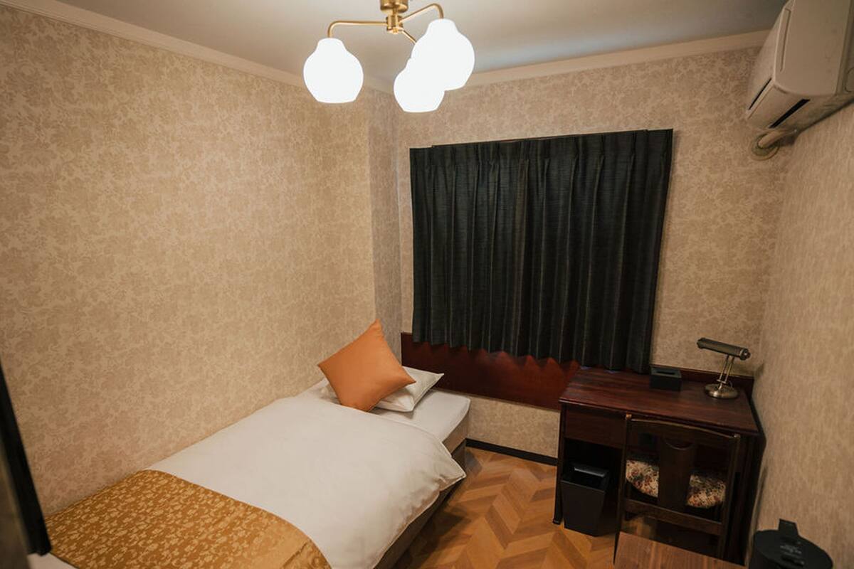 A hotel room with a bed, a desk, a chair, a small table, a lamp, and a window with curtains.