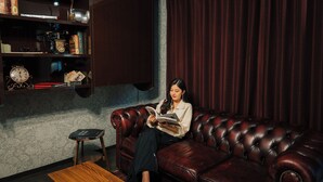 Una stanza con un divano in pelle rosso scuro, un piccolo tavolino in legno, una libreria con libri e oggetti decorativi, e una tenda scura.