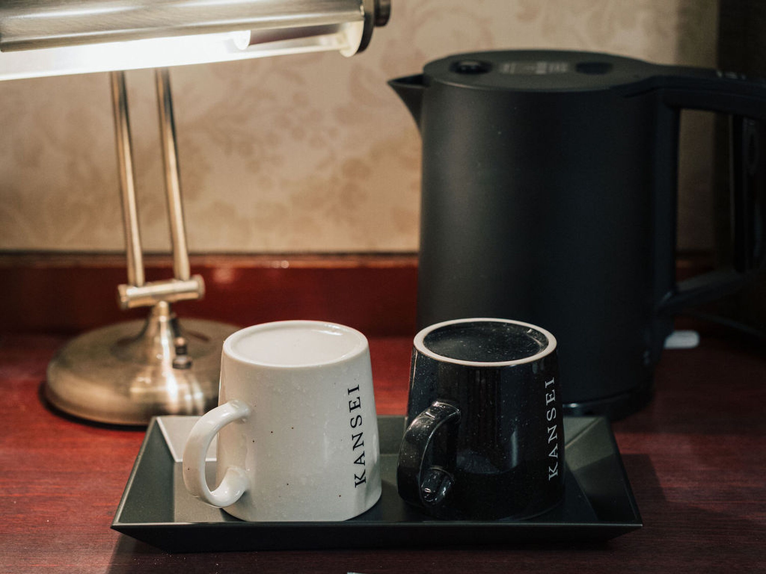 Una bandeja con una taza blanca, una taza negra, una cafetera y una lámpara sobre una mesa.