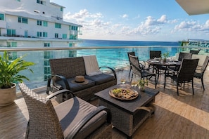 A balcony with wicker furniture, a dining table with a fruit platter, and a view of the ocean.