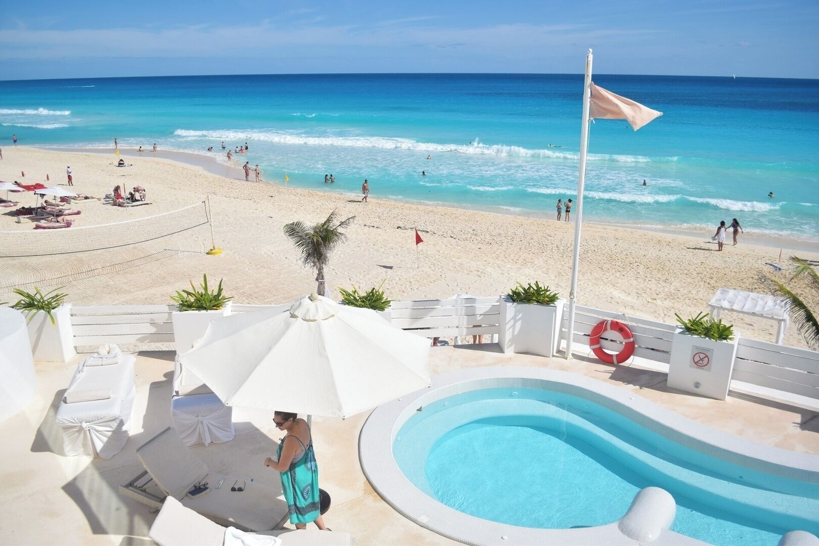 A pool area with lounge chairs and umbrellas, overlooking a beach with people and a lifebuoy.