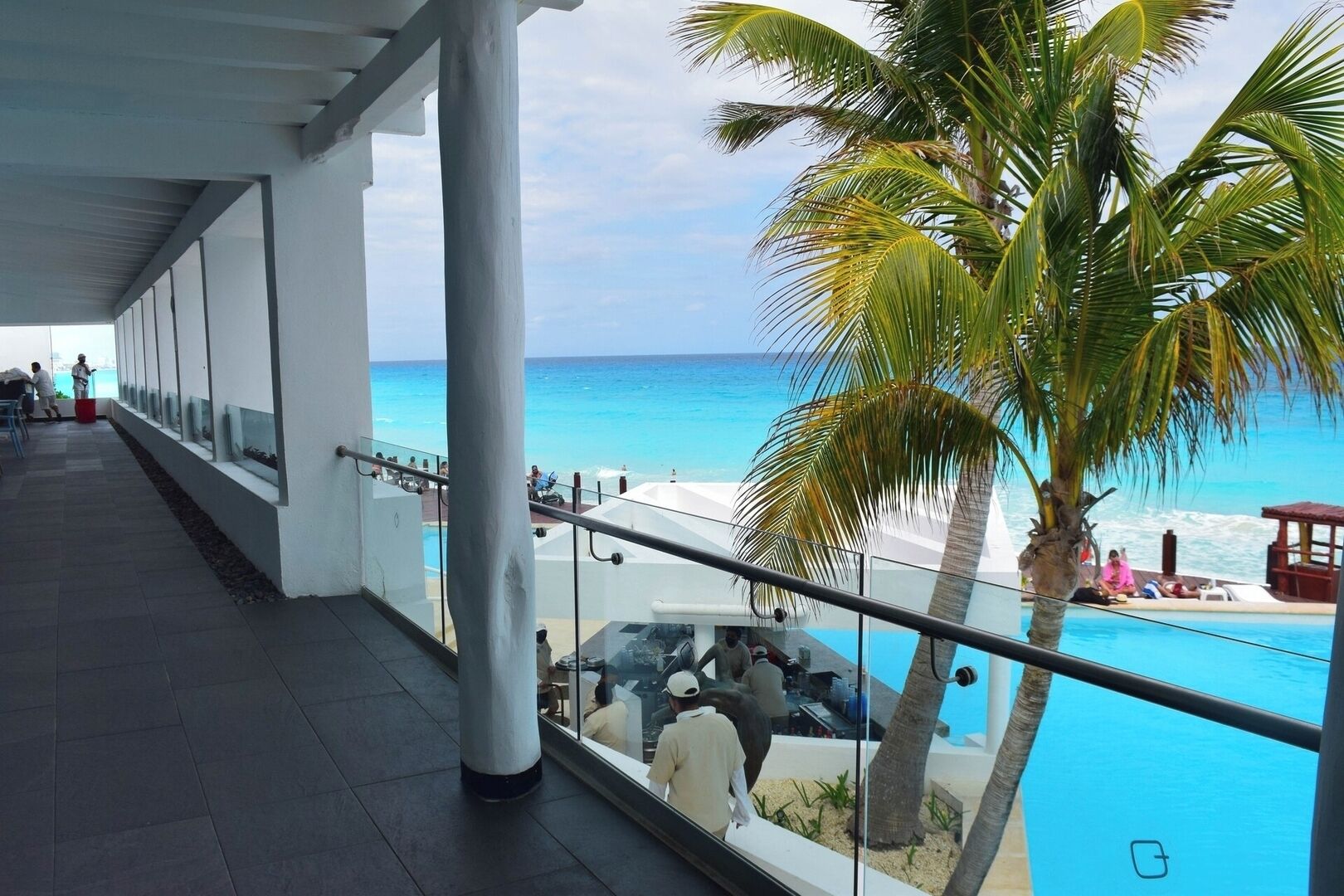 A balcony with a view of the ocean and a pool area with palm trees.