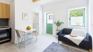 Une cuisine moderne dotée d’un four encastré, un coin repas avec une table et des chaises blanches, un canapé foncé agrémenté d’un plaid blanc, et une grande fenêtre donnant sur un paysage verdoyant.