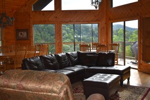 A wooden cabin interior with a large black leather sectional sofa, a dining table with chairs, and large windows offering a view of a forested mountain area.