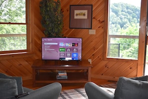 A wooden interior with a television on a stand, a framed picture on the wall, and a view of trees through the window. - 4 bedrooms and loft sleeps 12 with view  pool table 2 levels of decks and hot tub (Sevierville)