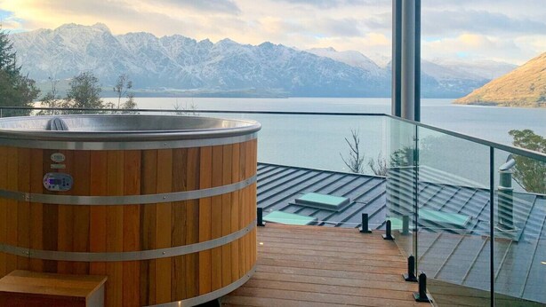 Uma banheira de hidromassagem de madeira em um deck de madeira com guarda-corpo de vidro, com vista para um lago e montanhas.