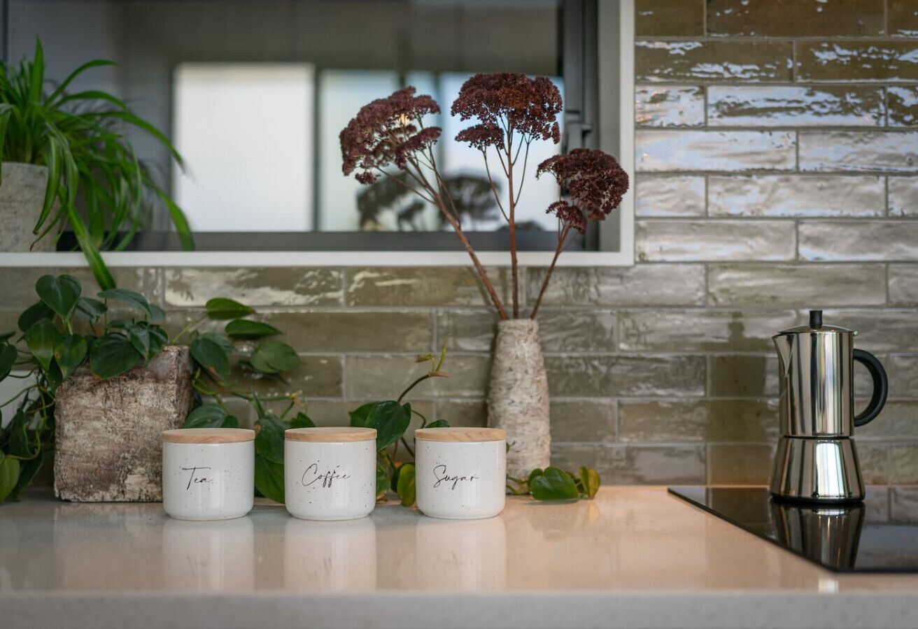 A kitchen counter with three white jars labeled "Tea," "Coffee," and "Sugar," a stainless steel coffee pot, and a vase with red flowers.