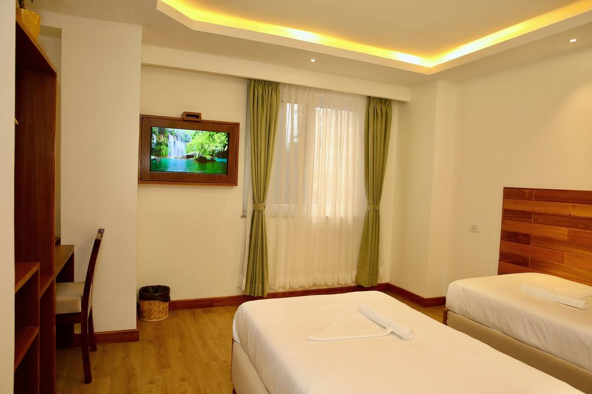 A hotel room with two beds, a TV mounted on the wall, a wooden headboard, and a window with sheer curtains.
