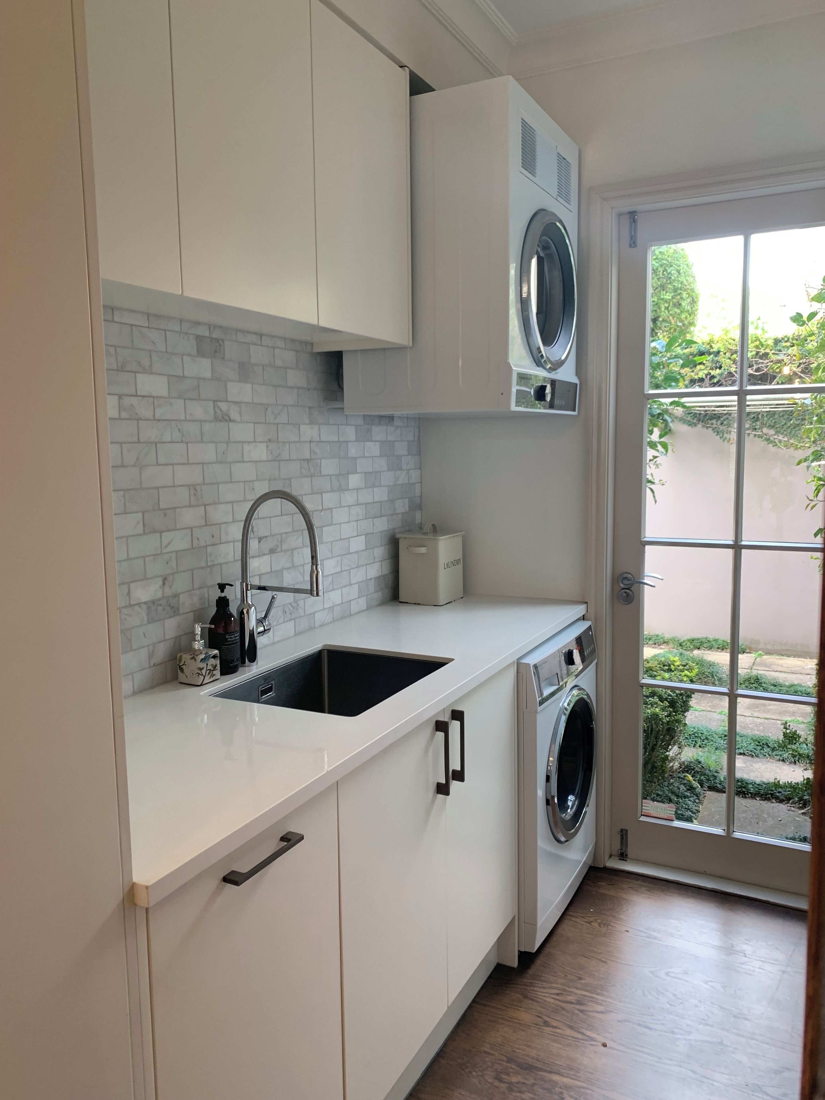 Une cuisine moderne avec des meubles blancs, un lave-linge encastré, un évier et une fenêtre donnant sur un espace verdoyant.