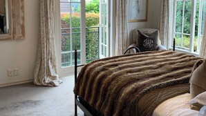 A bedroom with a large bed, a window with curtains, a mirror, and a view of greenery outside.