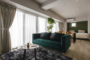 Un salon moderne avec un canapé vert, une petite table ronde, une plante en pot et un lit à l’arrière-plan.