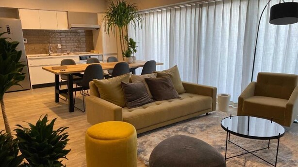 Un salon moderne avec un canapé, un pouf et une table basse, un coin repas avec des chaises, et une cuisine en arrière-plan.