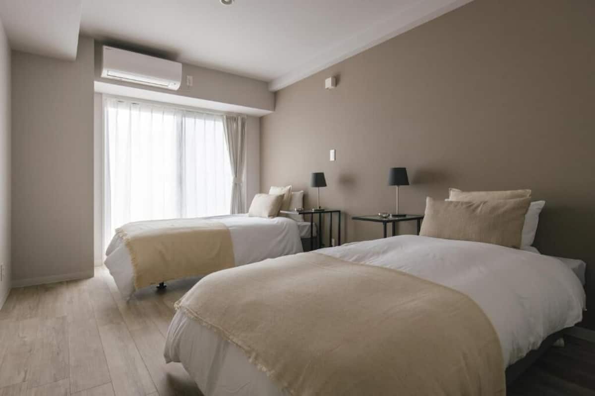A hotel room with two beds, an air conditioning unit, a window with curtains, and two bedside tables with lamps.