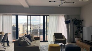 Un salon moderne comprenant un canapé, un pouf et un fauteuil, un coin repas avec des chaises, un téléviseur et une plante.