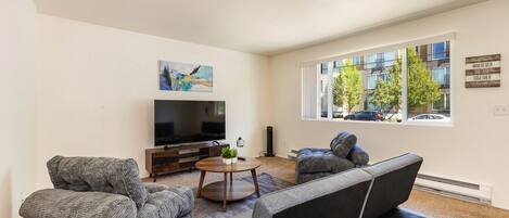 A modern living room with a large flat-screen TV, grey upholstered sofas, a coffee table, and a painting on the wall.