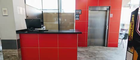A reception desk with a red tiled wall, a closed elevator, and a sign indicating the hotel name.