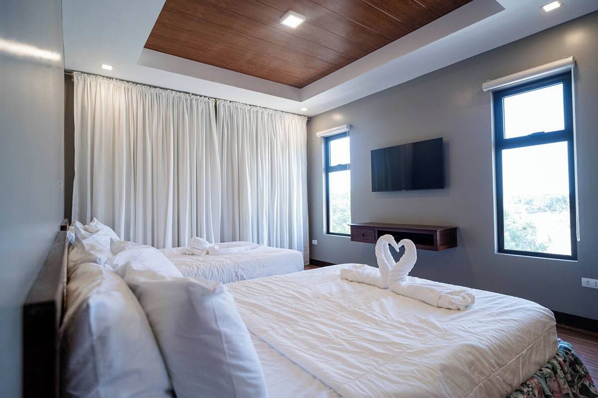A hotel room with two beds, a flat-screen TV mounted on the wall, and large windows with curtains.
