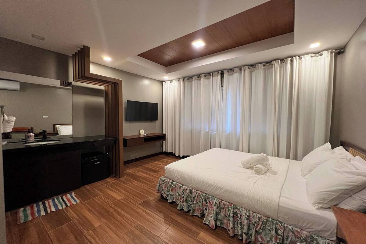 A modern hotel room with a wooden floor, a bed with white linens, a built-in desk with a flat-screen TV, and a large window with curtains.