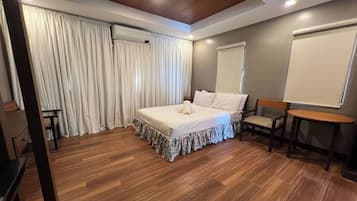 A bedroom with a wooden floor, a bed with a floral bed skirt, a wooden chair, a small table, and a whiteboard on the wall.
