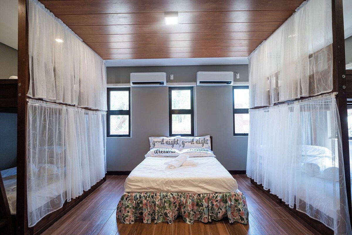 A bedroom with a wooden ceiling, air conditioning units, a bed with floral bedding, and sheer curtains.