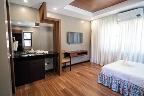 A hotel room with a wooden floor, a large mirror, a flat-screen TV, a bed with a floral skirt, and a desk with a chair. - Villa Minerva Eco-farm Resort Near Vigan By Zuzu (Caoayan)