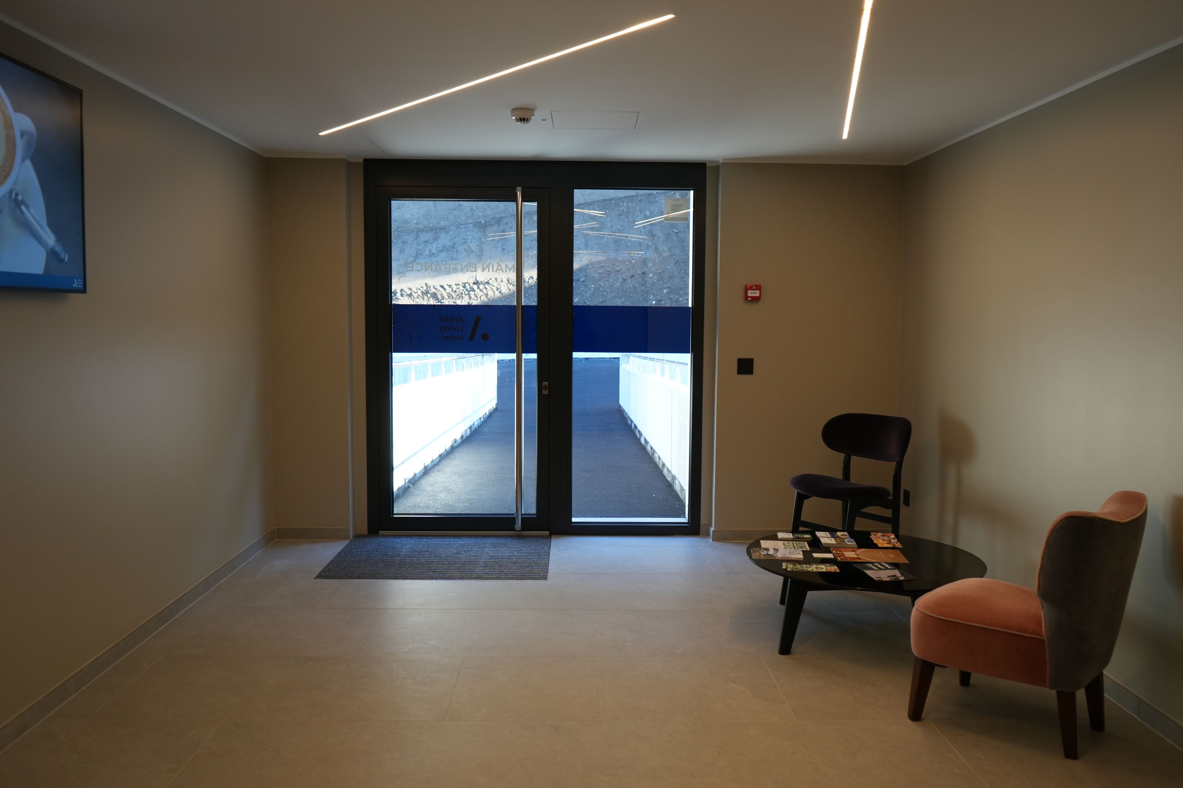 Un hall d’hôtel avec une porte vitrée donnant sur un couloir, un comptoir d’accueil et un espace détente avec deux fauteuils.