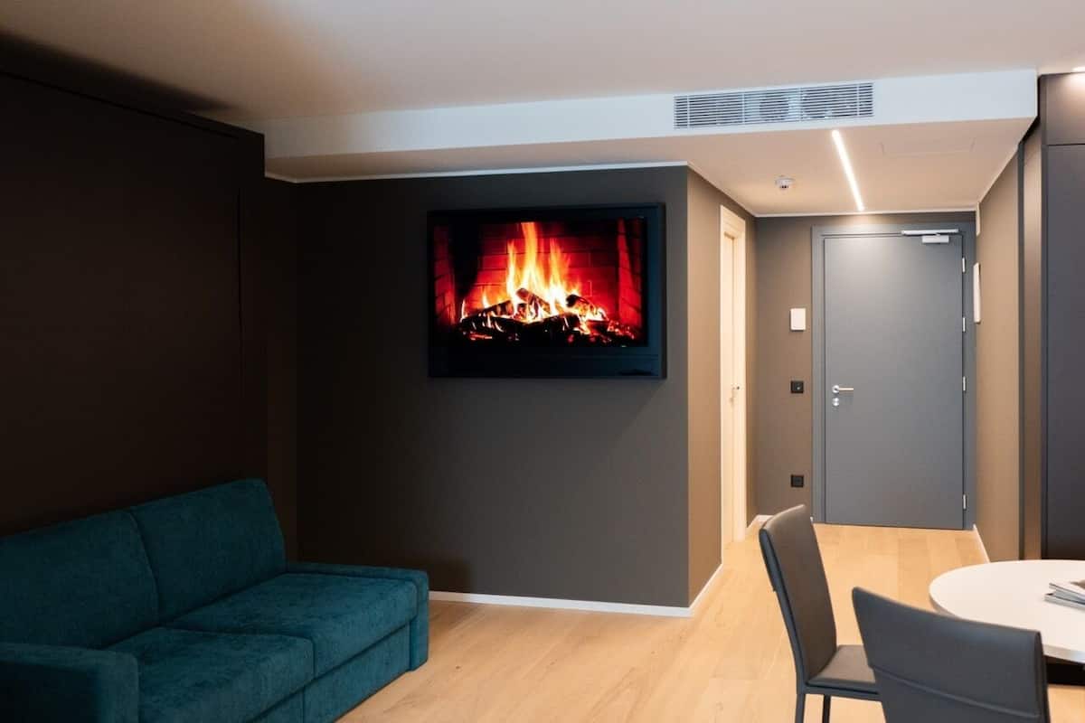 A modern living room with a flat-screen TV displaying a fireplace, a teal sofa, and a dining area with chairs.