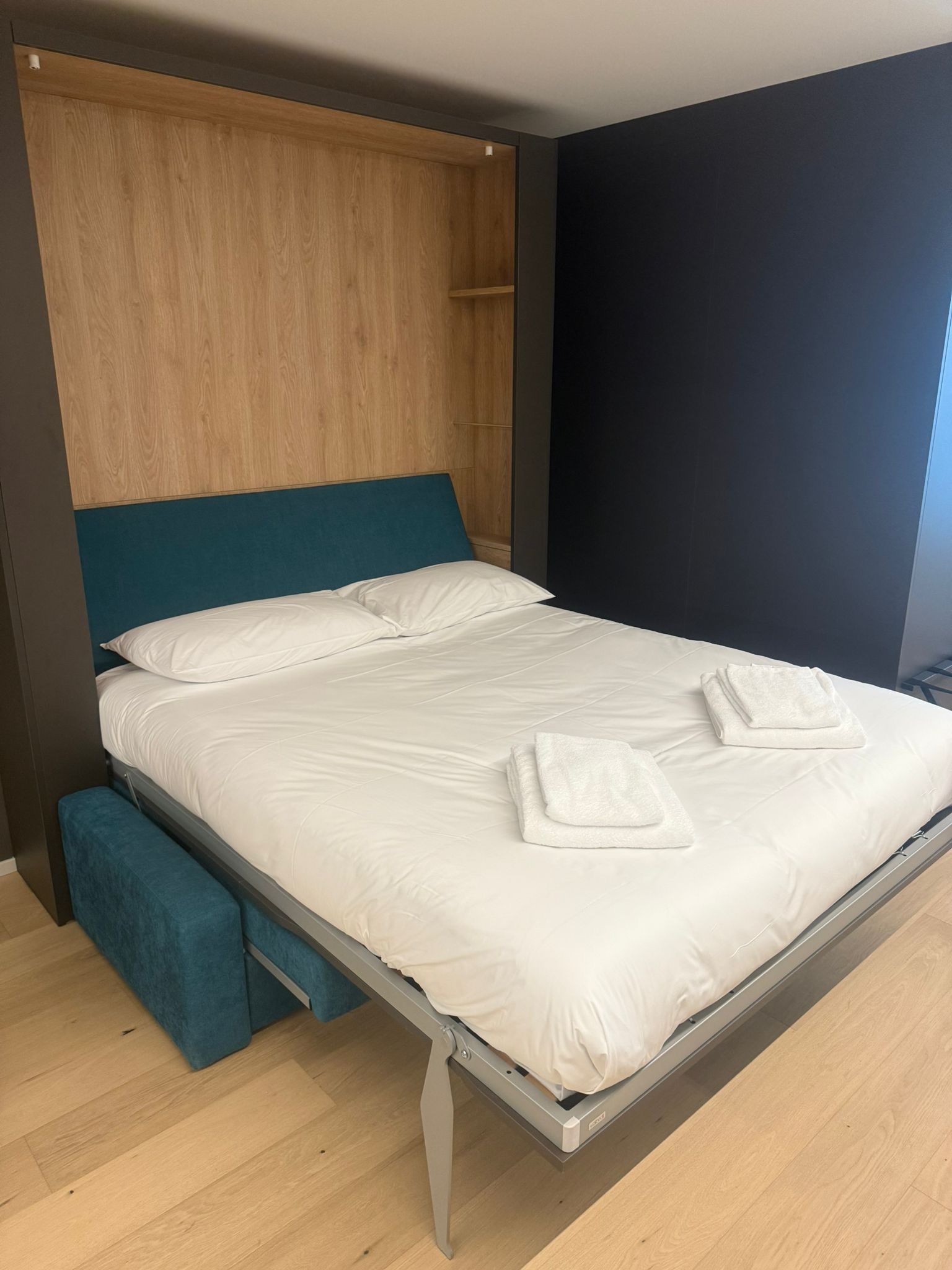 Un lit moderne avec une tête de lit en bois, un banc au pied du lit et un mur foncé sur le côté droit.