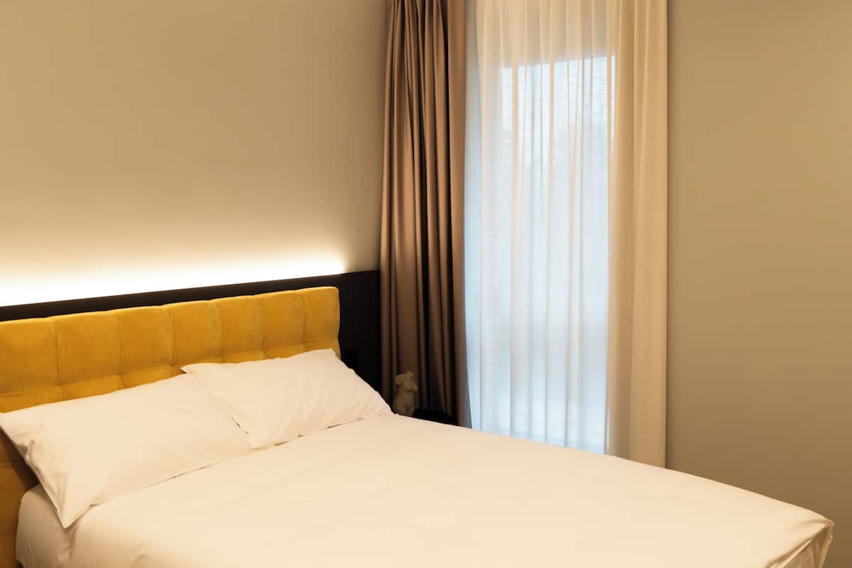 A neatly made bed with white linens and a yellow headboard, a window with sheer curtains, and a wall-mounted light fixture.