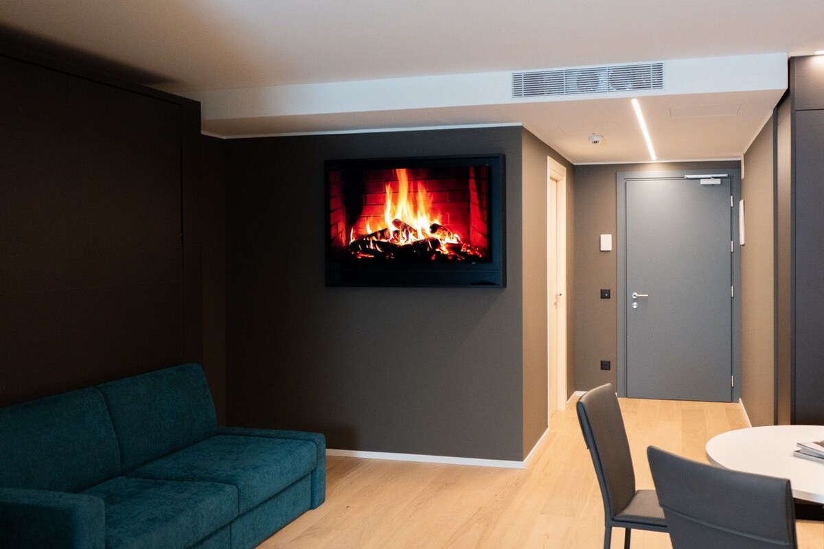 A modern living room with a flat-screen TV displaying a fireplace, a teal sofa, and a dining area with chairs.