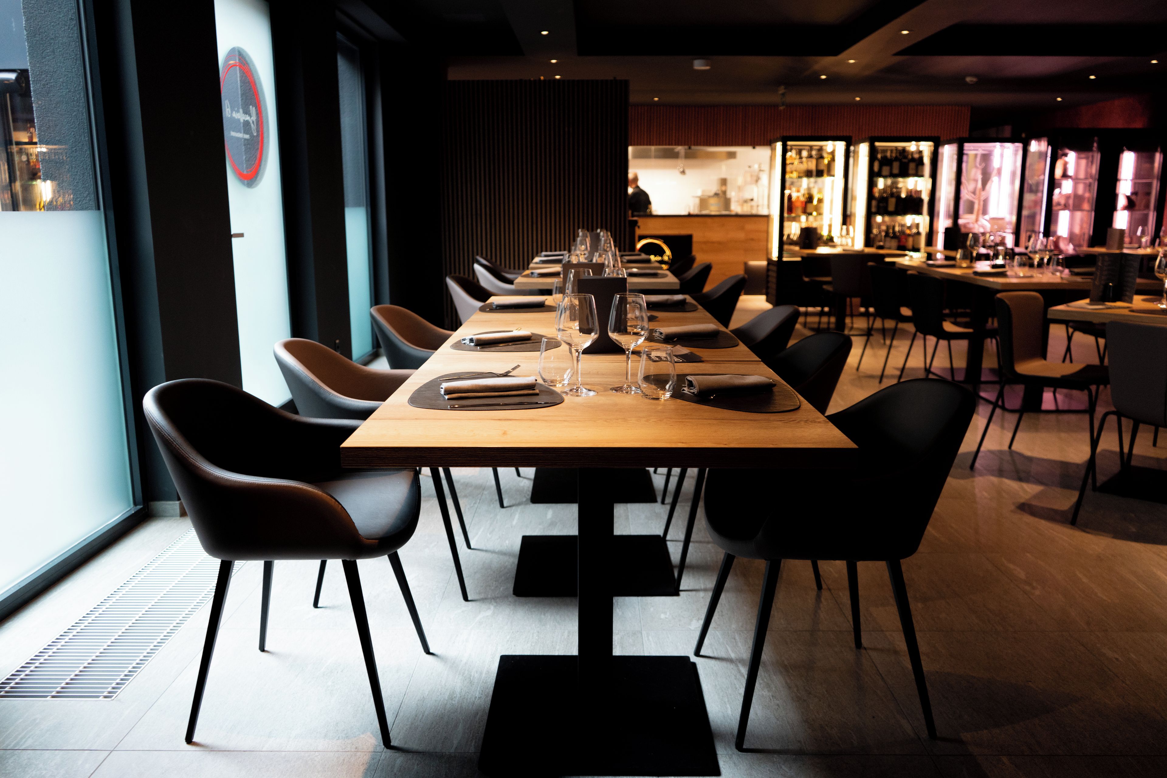 Un restaurant moderne avec une longue table en bois dressée pour le dîner, des chaises noires et un bar situé au fond de la salle.
