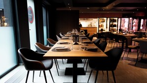 A modern restaurant with a long wooden table set for dining, black chairs, and a bar area in the background. - Arbed Living Hotel Self Check-in (Arbedo-Castione)