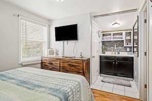 A bedroom with a bed, a TV, a wooden dresser, and a bathroom with a sink and mirror. - Tiki Retreat-Hot Tub-Fire Pit - Private Beach (Sand Lake)
