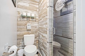 A bathroom with a wooden wall design, a toilet, a glass shower enclosure, and a sign reading 'Blue Lagoon Paradise.' - Tiki Retreat-Hot Tub-Fire Pit - Private Beach (Sand Lake)