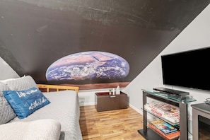 A living room with a large flat-screen TV, a sofa, a coffee table, and a bookshelf with magazines.