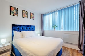 A bedroom with a large bed, a nightstand, a window with blinds, and three wall-mounted artworks.