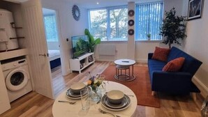 A modern living room with a blue sofa, a dining table set for two, and a large window with blinds.