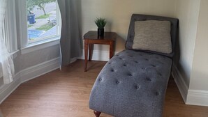 A bedroom with a grey upholstered bed, a small wooden nightstand with a plant, and a window with light curtains. - Cute 3BR Upper Unit Home in South Buffalo near downtown (Buffalo)