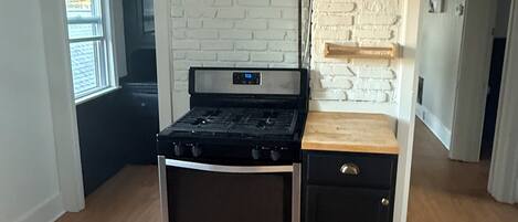 A compact kitchen with a built-in stove and oven, white brick walls, and a wooden countertop.