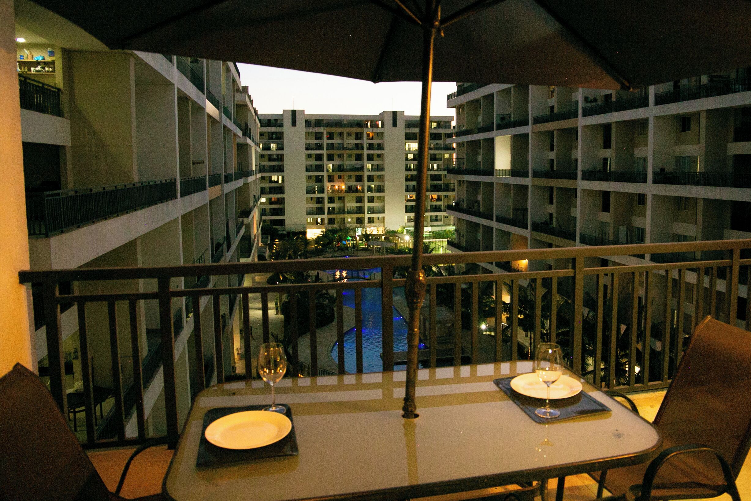 A balcony with a table set for two, overlooking a swimming pool and a high-rise building.